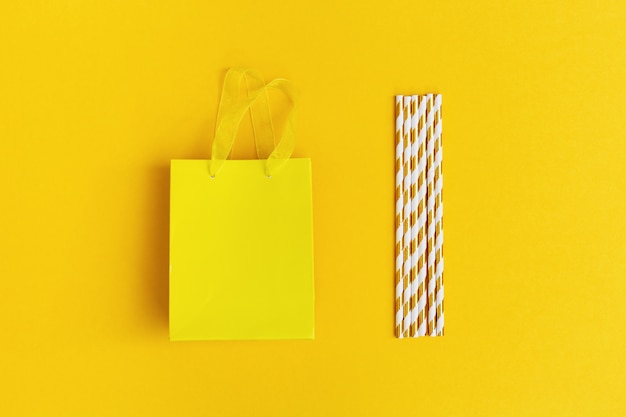 Bolsa de papel de regalo y pajitas con rayas doradas sobre fondo amarillo. Concepto de fiesta, fiesta, cumpleaños. Imagen sin sombra con espacio de copia para su texto. Vista superior y endecha plana.