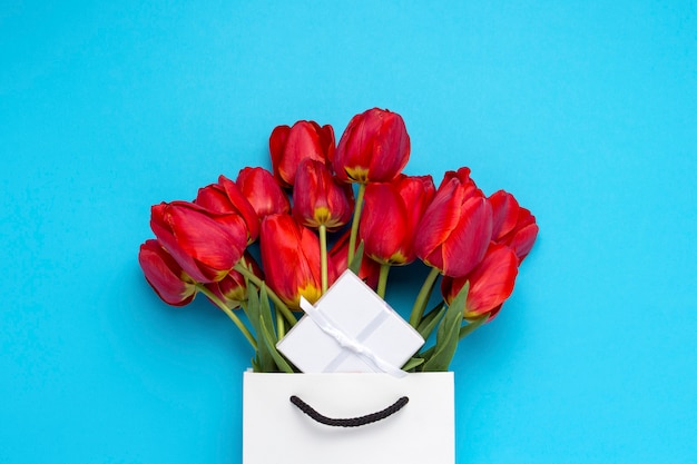 Bolsa de regalo blanca, una pequeña caja de regalo blanca y un ramo de tulipanes rojos sobre un azul. Concepto Ofrece un compromiso o matrimonio, ir de compras