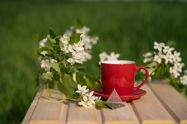 una bolsita de té desechable junto a una taza roja y una rama de manzano en flor en una mesa de madera