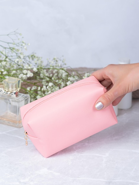 Bolso cosmético rosa de cuero sobre una mesa de piedra con flores y perfume en el fondo