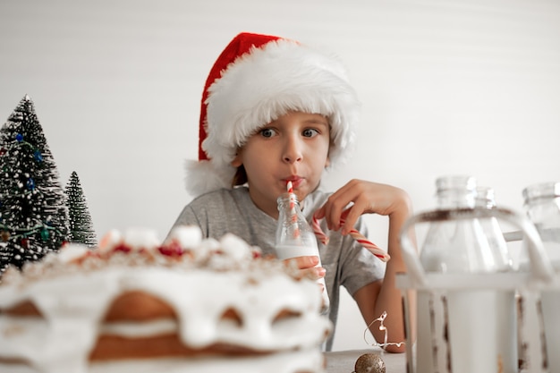 Foto bom dia de natal. um menino loiro com um chapéu de papai noel vermelho está tomando café da manhã, bebendo leite.