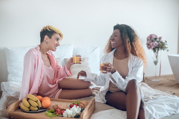 Bom Dia. Duas mulheres bonitas rindo comendo na cama no quarto relaxadas e alegres