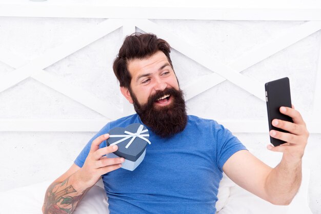 Bom dia Presente especial Homem barbudo usando tecnologia móvel na cama Cara bonito tomando selfie com caixa de presente Selfie feliz Vida moderna nova tecnologia Conceito de tecnologia Conversa agradável