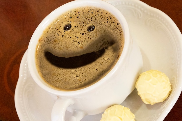 Bom dia, xícara de café branca com um sorriso e doces em um fundo de madeira, close-up, conceito - bom humor