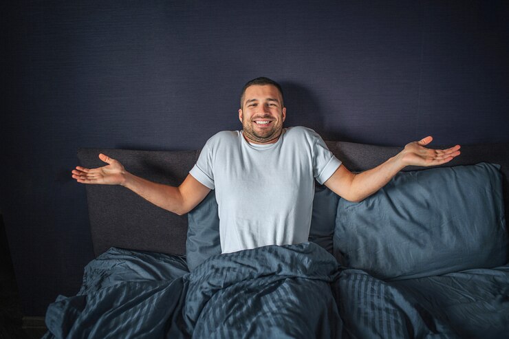 Foto bom e positivo jovem sentado na cama. ele está coberto com um cobertor azul escuro. ele mantém as mãos afastadas do corpo. cara sorri. ele está feliz.