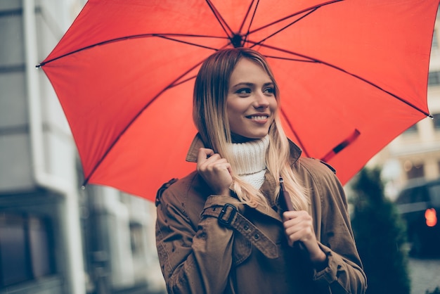 Bom humor em qualquer clima. Mulher jovem e atraente sorridente carregando guarda-chuva e ajustando o casaco enquanto caminha pela rua