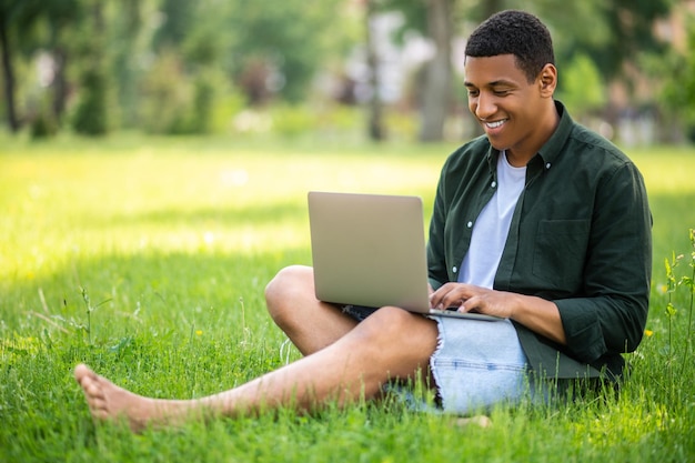 Bom humor. Feliz cara de pele escura descalço digitando no laptop sentado na grama na natureza em dia ensolarado