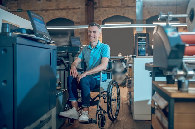 Bom humor. homem adulto confiante sorridente em uma camiseta azul e calça jeans em uma cadeira de rodas perto de equipamento de escritório trabalhando em um espaço de escritório