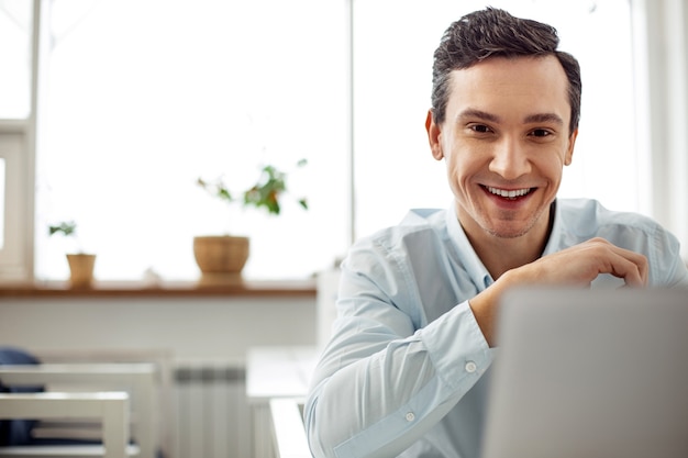 Bom humor. Homem de cabelos escuros, olhos escuros, alerta atraente, sorrindo e trabalhando em seu laptop enquanto está sentado à mesa