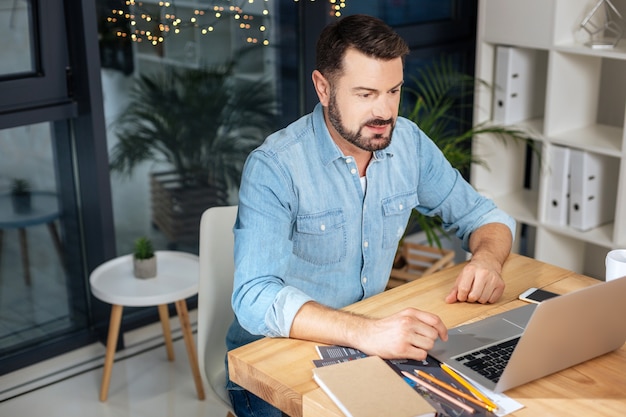 Bom humor. Homem simpático, positivo e bonito olhando para a tela do laptop e sorrindo enquanto está de bom humor