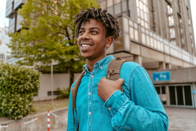 Foto bom humor. jovem de pele escura com mochila sorrindo andando na rua da cidade perto do prédio de escritórios em um belo dia