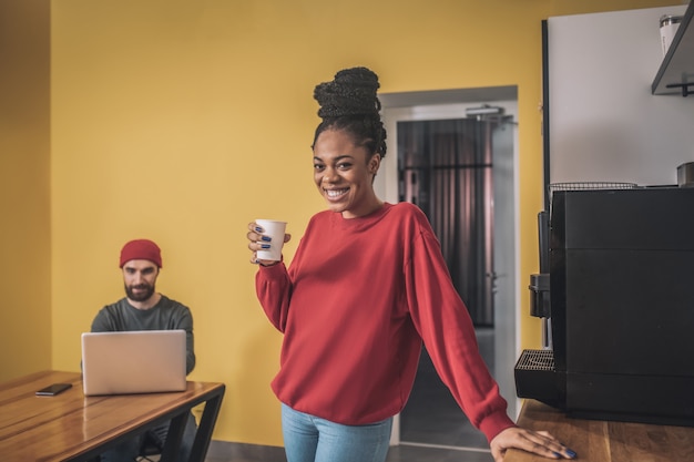 Bom humor. Menina bonita alegre de pele escura tomando café e um cara barbudo com um laptop sentado à mesa atrás do escritório