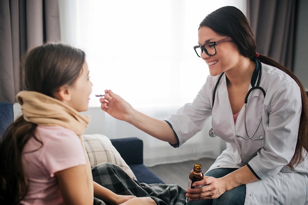 Bom jovem médico feminino sorrindo. ela dá xarope na colher para criança doente. médico direcioná-lo para a boca do garoto. menina pequena, mantenha-a aberta. eles estão em um quarto.