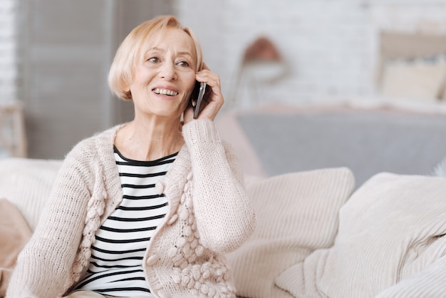 Bom ouvir você. Mulher simpática e bonita usando seu telefone para falar com alguém enquanto está sentada em um sofá