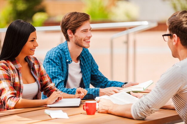 Bom tempo com amigos da faculdade. Grupo de jovens alegres, sorrindo e discutindo algo enquanto estão sentados na mesa de madeira ao ar livre