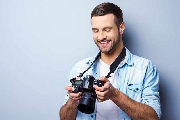 Bom tiro! Jovem bonito segurando uma câmera digital e olhando para ela com um sorriso em pé contra um fundo cinza