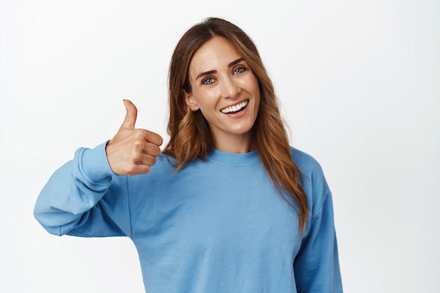 Bom trabalho. Mulher morena sorridente e satisfeita mostrando o polegar em aprovação, aceno, elogio, ótimo trabalho, satisfeita com a qualidade, recomendando, pisando em branco