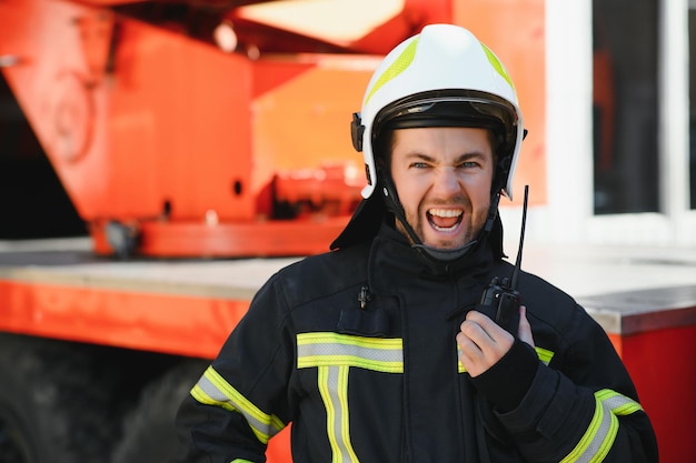 Foto bombeiro em um uniforme de proteção ao lado de um caminhão de bombeiros e falando no rádio