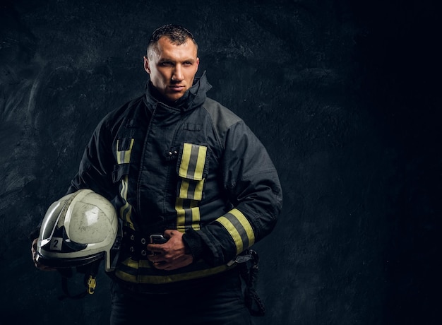 Bombero confiado en uniforme sosteniendo un casco y mirando a la cámara, de pie en el estudio contra una pared de textura oscura