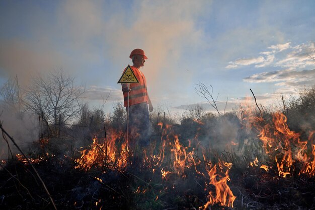 Bombero ecologista luchando contra el fuego en el campo por la noche Hombre con chaleco y casco cerca de la hierba ardiente con humo sosteniendo un triángulo amarillo con cráneo y huesos cruzados signo de advertencia Concepto de desastre natural