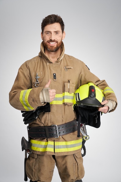 Bombero profesional de pelo oscuro manteniendo el pulgar hacia arriba y sonriendo en el retrato de estudio de un alegre barbudo
