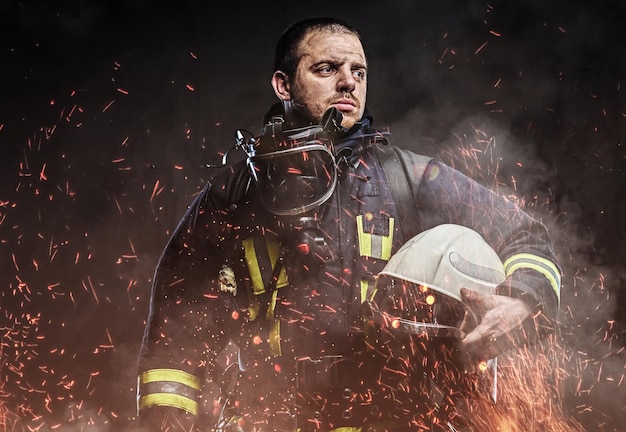 Un bombero profesional vestido con uniforme con casco de seguridad en chispas de fuego y humo sobre un fondo oscuro.