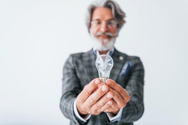 Con bombilla en las manos Senior elegante hombre moderno con pelo gris y barba en el interior