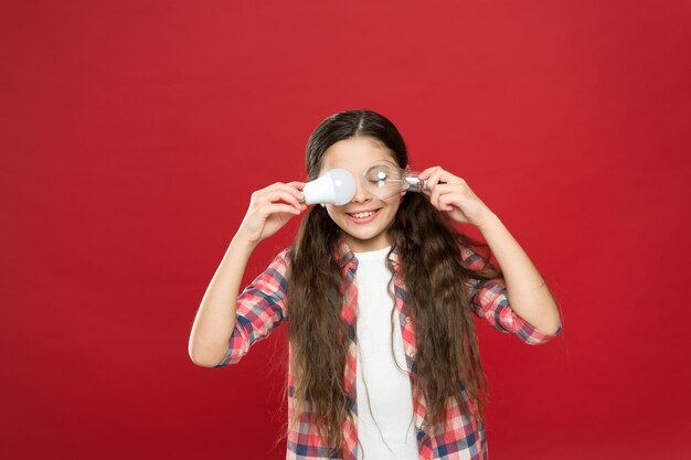 Bombilla de niña pequeña. energía y electricidad. creando una gran idea. Pensando en la niña. niño pequeño sostenga la bombilla de la idea de luz. concepto de educación. niño de escuela pequeño fondo rojo.