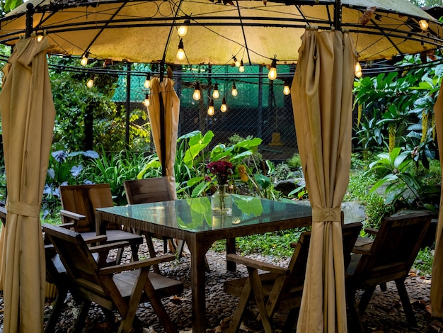 Bombillas antiguas colgadas en la vieja carpa sobre la mesa de comedor vacía, mesa de vidrio y sillas de madera en la escena nocturna del jardín al aire libre sin gente