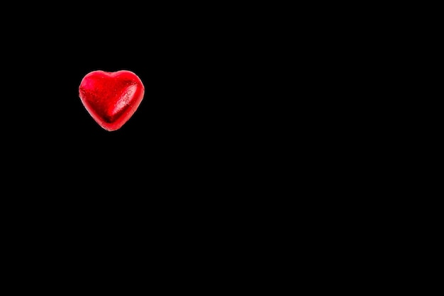 Bombom de chocolate em forma de coração envolto em papel filme vermelho no lado esquerdo, sobre fundo preto.