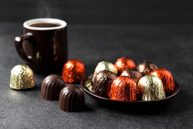 Bombons de chocolate embrulhados em papel alumínio multicolorido em um prato e uma xícara de café quente na superfície preta com espaço de cópia