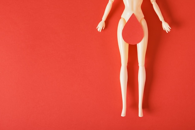 Boneca com uma gota de sangue de papel nas coxas, arte feminista, conceito de saúde e gnecologia da mulher, espaço de cópia de fundo vermelho menstruação