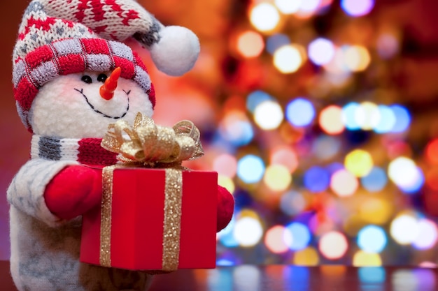 Boneco de neve de Natal e ano novo com caixa de presente vermelha no fundo de luzes desfocadas da árvore de Natal. copie o espaço.