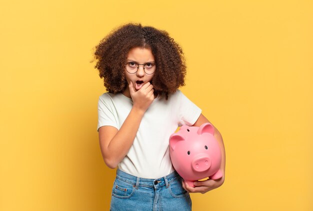 Bonita adolescente afro cubriéndose la boca con las manos con una expresión de asombro y sorpresa, guardando un secreto o diciendo uy. concepto de ahorro