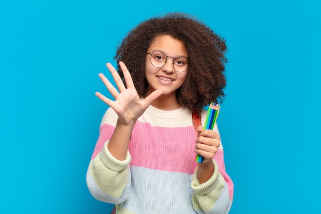 Bonita adolescente afro sonriendo y mirando amigable, mostrando el número cinco o quinto con la mano hacia adelante, contando hacia atrás. concepto de estudiante