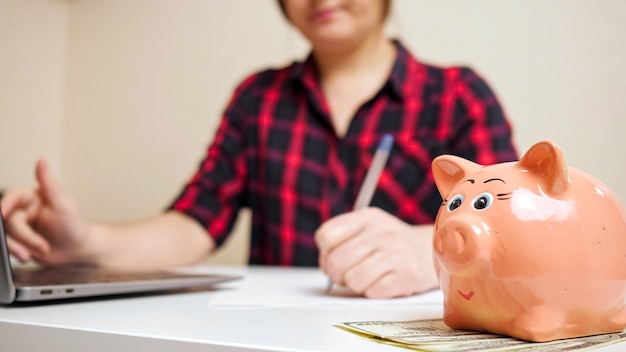 Bonita alcancía rosa se encuentra en billetes de dólar contra la señora borrosa escribiendo en el teclado de la computadora portátil y contando los gastos del niño vista cercana