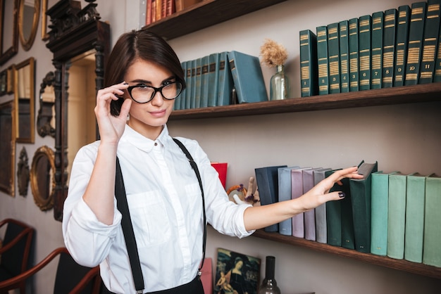 Bonita autora de gafas y camisa blanca seleccionando el libro en la estantería