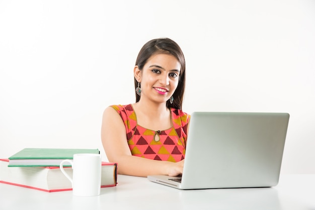 Bonita chica asiática india estudiando en un ordenador portátil con un montón de libros sobre la mesa, sobre fondo blanco.