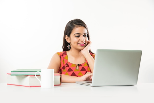 Bonita chica asiática india estudiando en un ordenador portátil con un montón de libros sobre la mesa, sobre fondo blanco.