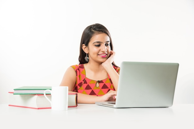 Bonita chica asiática india estudiando en un ordenador portátil con un montón de libros sobre la mesa, sobre fondo blanco.