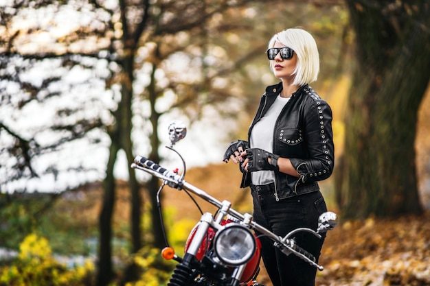 Bonita chica biker rubia en gafas de sol con moto roja en la carretera en el bosque