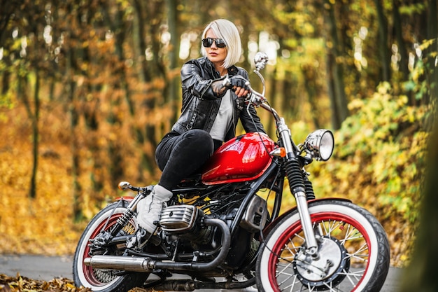 Bonita chica biker rubia en gafas de sol con moto roja en la carretera en el bosque