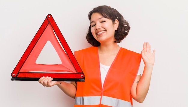 Bonita chica hispana sonriendo felizmente agitando la mano dándole la bienvenida y saludando el triángulo del accidente automovilístico