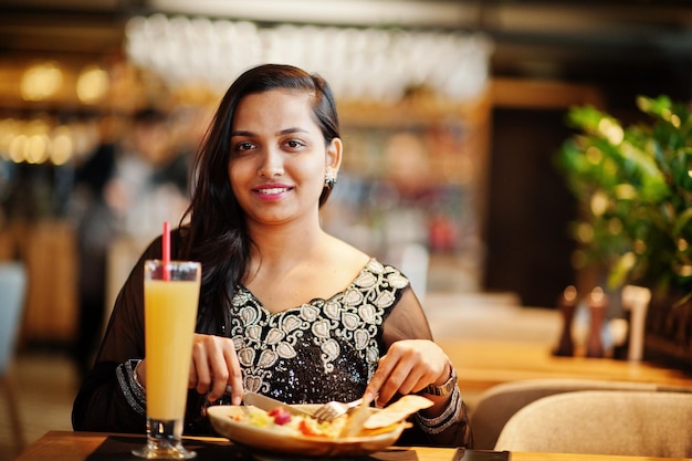 Bonita chica india con vestido de sari negro posó en el restaurante sentado a la mesa con jugo y ensalada