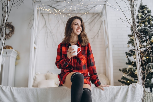 Bonita chica morena en la cama con camisa roja y calcetines calientes con taza de café y luces de Navidad detrás