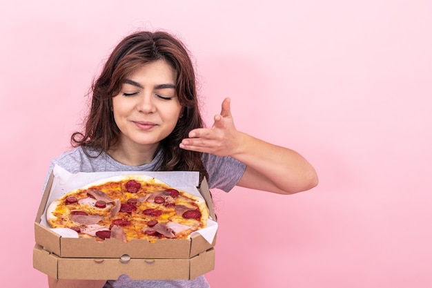 Una bonita chica morena disfruta del olor de la pizza recién hecha de una caja de entrega sobre un fondo rosa, copie el espacio.