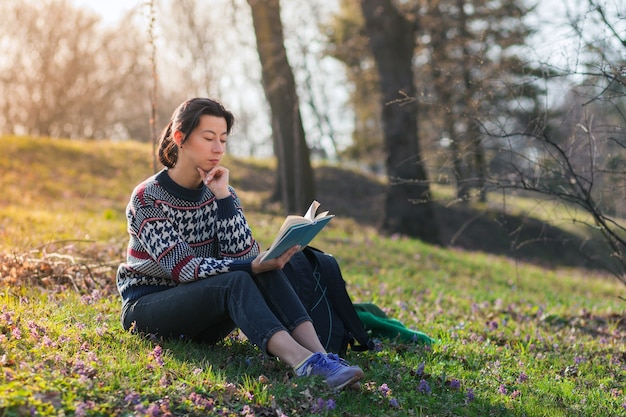 Bonita chica morena está sentada en el césped del parque y leyendo un libro.