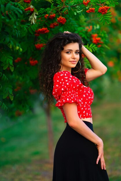 Bonita chica morena rizada con traje gitano rojo y accesorios posando en la naturaleza con serba