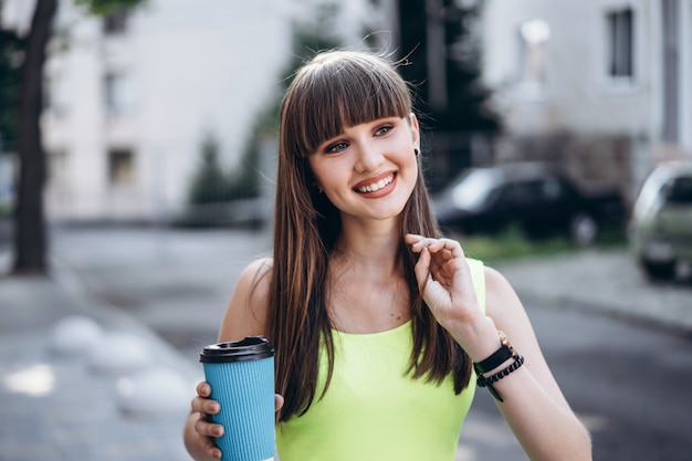 Bonita chica morena vestida de verde con una taza de café caminando al aire libre en la calle