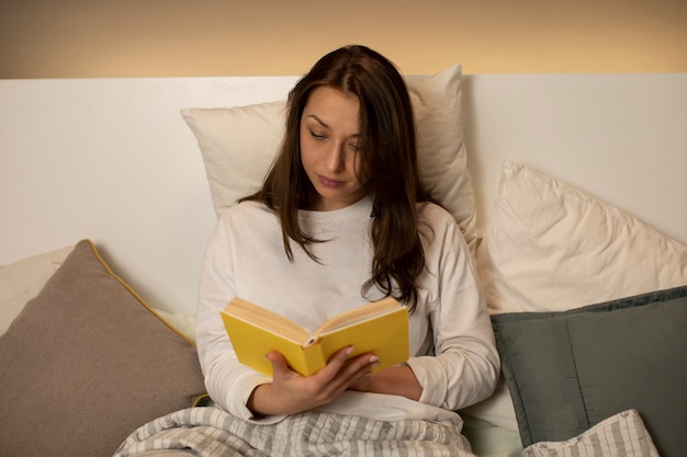Bonita chica de pelo oscuro en pijama leyendo libro con tapa amarilla sentado en la cama